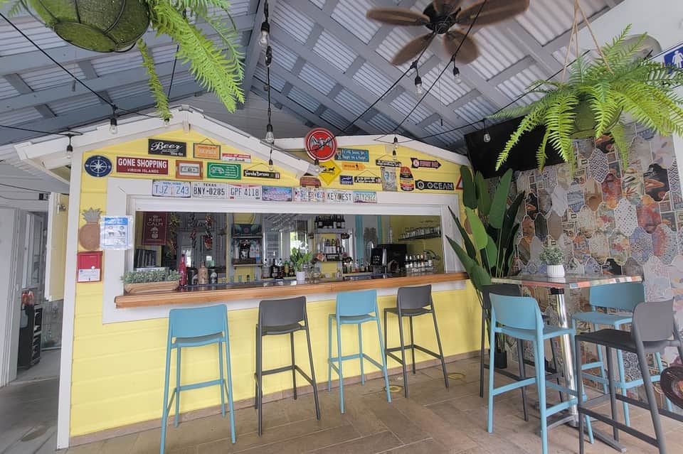 Outdoor bar with yellow storefront, blue stools, tropical plants, and casual seating area