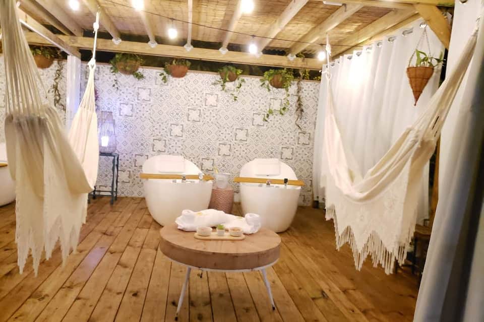 Spa treatment room with two white soaking tubs, wooden beams, hanging plants, and cream drapes