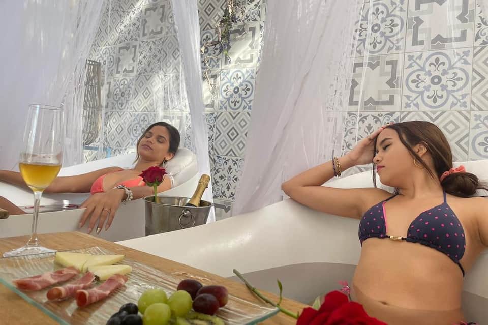 Two women relaxing in white bathtub with decorative tile backdrop and champagne service