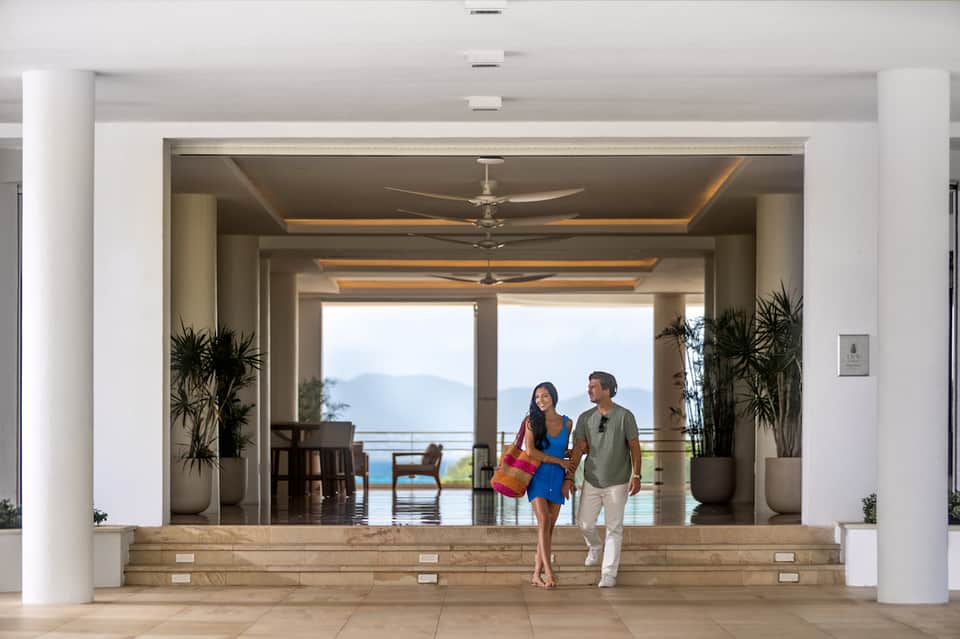 Bright modern lobby with white columns, potted plants, ceiling fans, and two people walking toward an ocean view terrace
