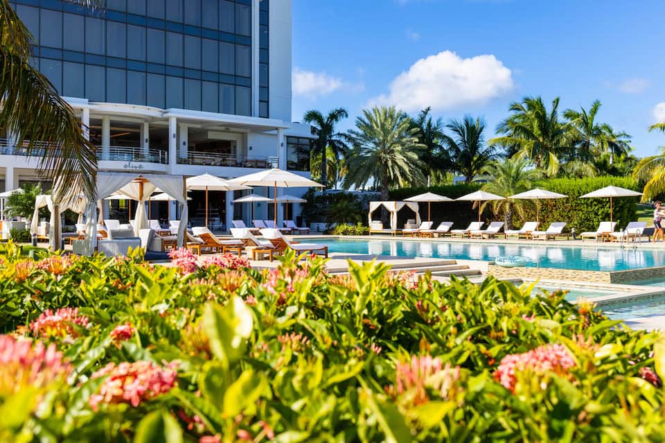 Resort pool area with loungers, umbrellas, and modern hotel building surrounded by tropical landscaping