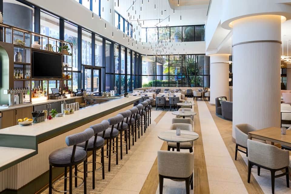 Modern hotel lobby bar with long counter seating, floor-to-ceiling windows, and contemporary décor