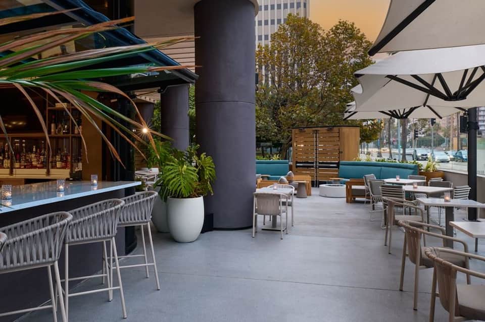 Modern rooftop bar and lounge with waterfront views, white wicker seating, blue umbrellas, and potted plants