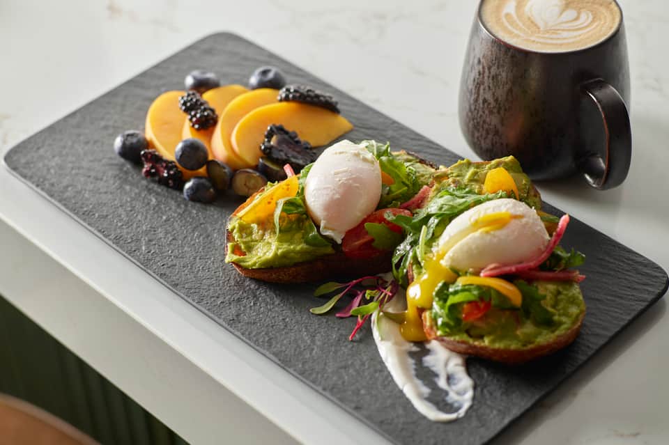 Gourmet breakfast plate with avocado toast, poached eggs, fresh fruit, and latte on dark slate