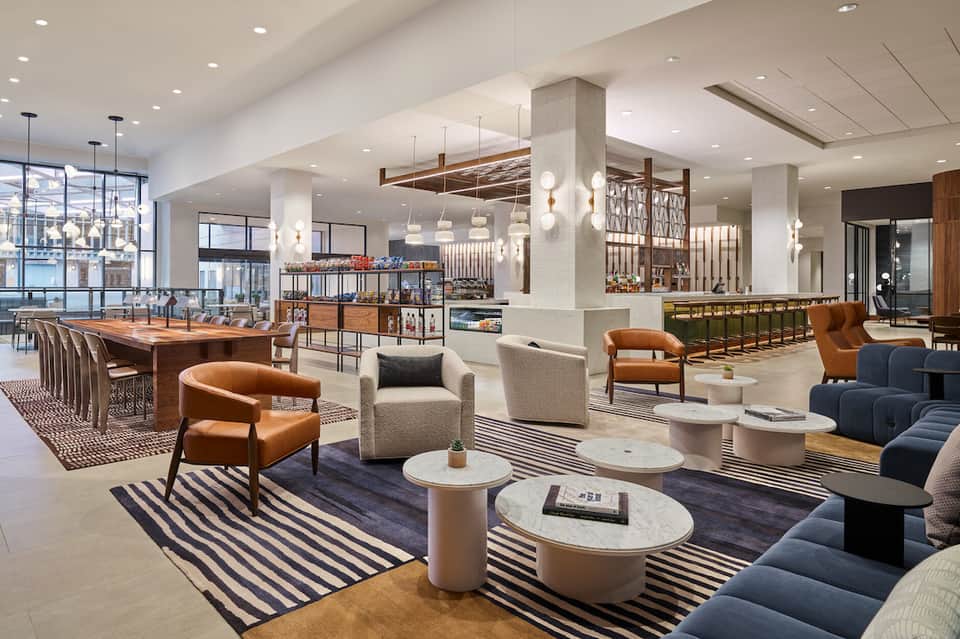 Modern hotel lobby with wood and marble finishes, orange leather chairs, pendant lights, and striped rugs