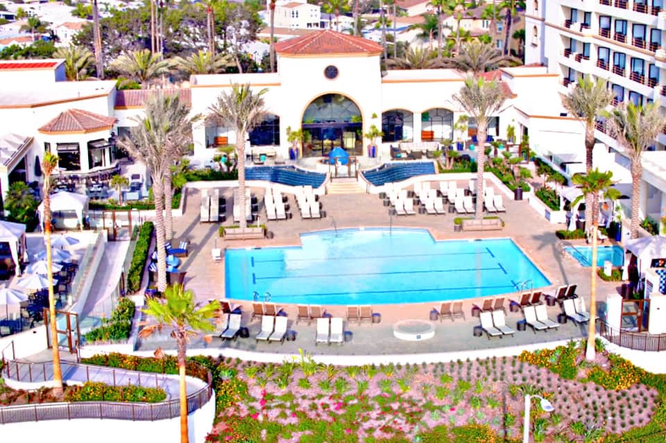 Aerial view of resort with rectangular swimming pool, white lounge chairs, palm trees, and Spanish-style central building