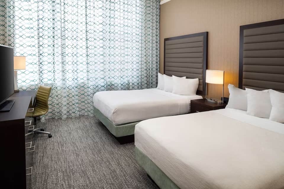 Double queen hotel room with patterned curtains, bedside lamps, and work desk