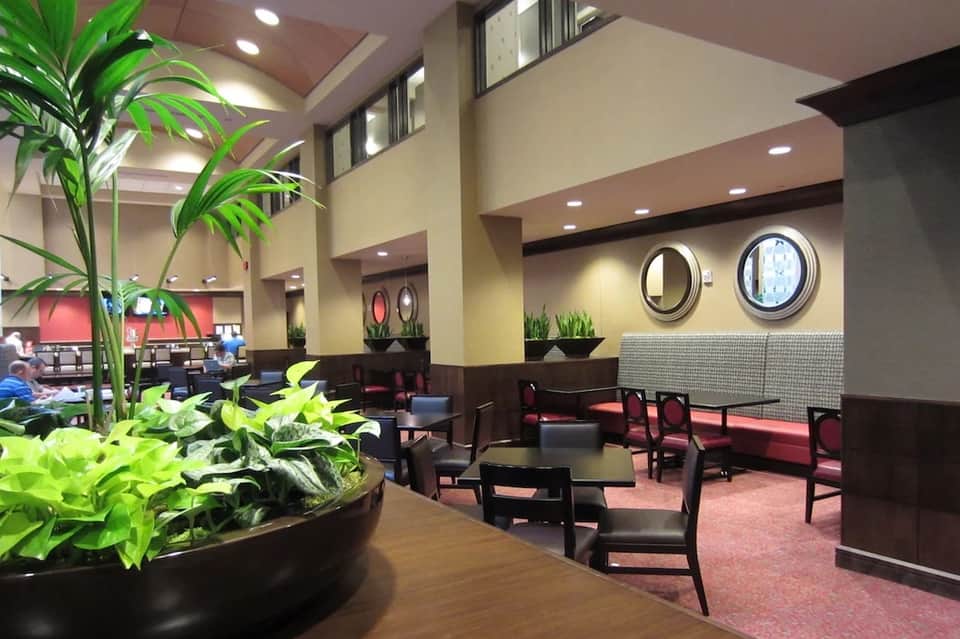 Hotel lobby lounge with high ceilings, potted plants, round mirrors, dark wood furniture, and soft lighting