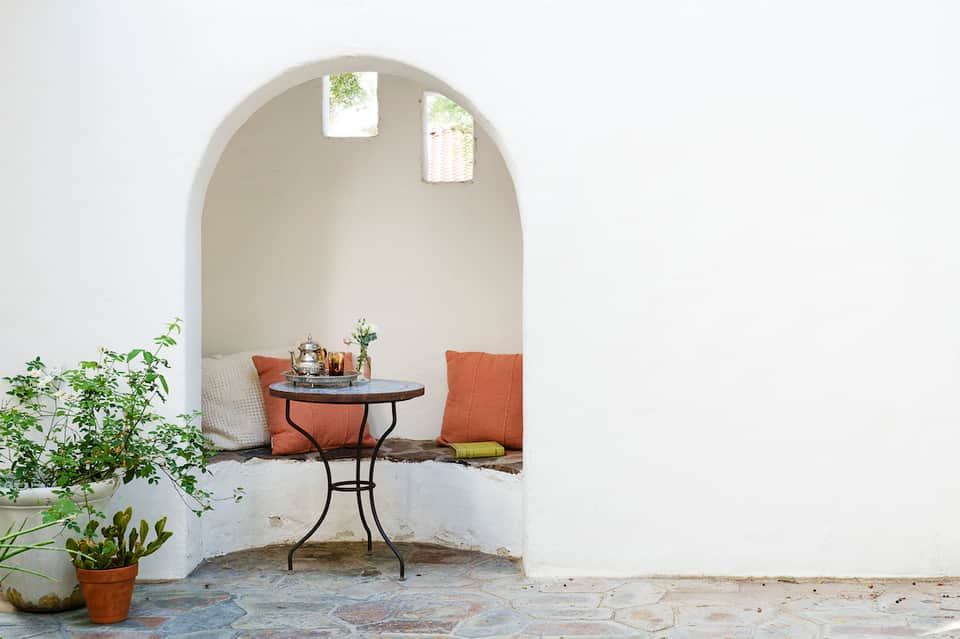 Mediterranean-style alcove with white arched entrance, orange cushioned chairs, small table, and potted plants