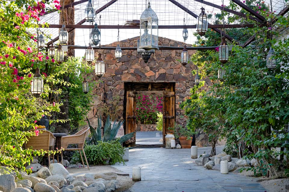 Desert courtyard with stone archway, wooden doors, climbing flowers, hanging lanterns, and decorative boulders