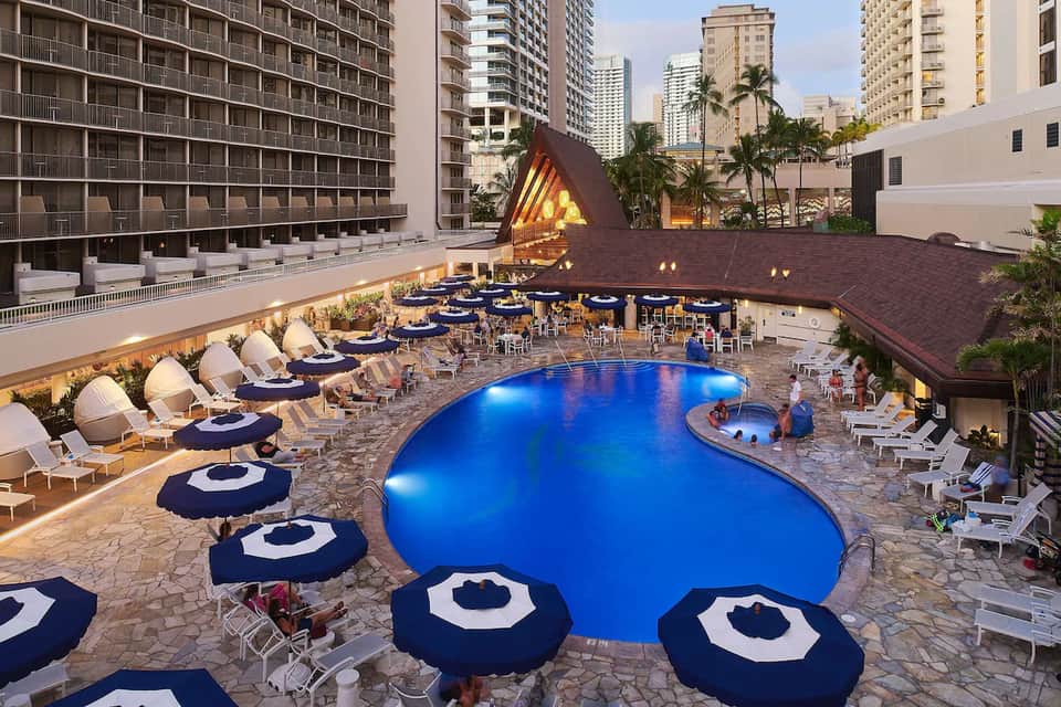 Illuminated resort pool at dusk with blue umbrellas, lounge seating, and high-rise hotel buildings