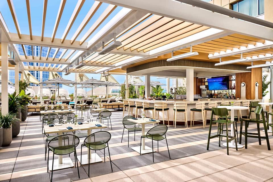 Modern rooftop bar with retractable pergola, lounge seating, green chairs, bar counter, and city skyline views