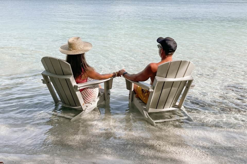 Two people in Adirondack chairs holding hands in shallow ocean water at the beach