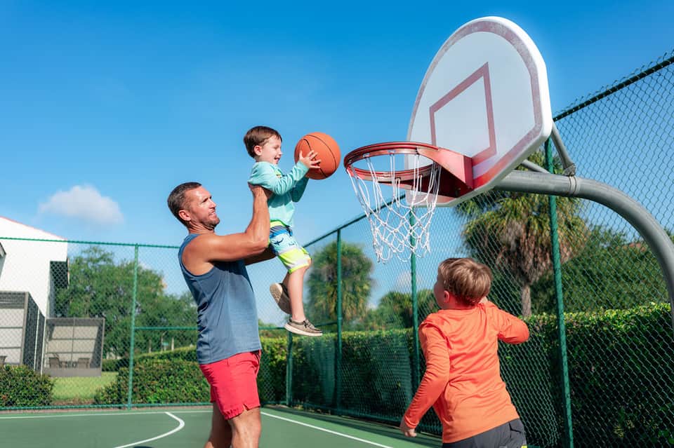Father lifting toddler to shoot basketball while child watches on outdoor court with fence