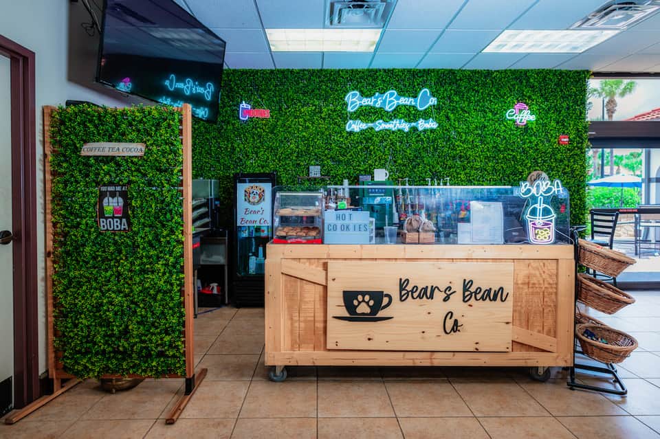 Coffee shop counter with wooden signage, green plant wall backdrop, and neon blue 'Bear's Bean Co' branding