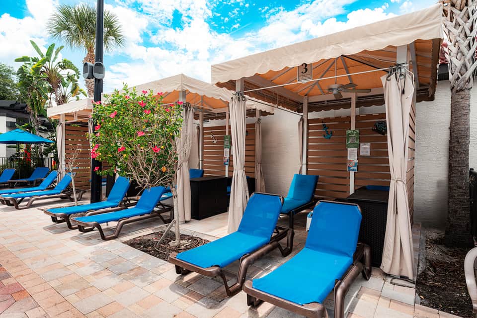 Outdoor cabana lounge area with blue chaise lounges, orange umbrellas, and pink flowering plants