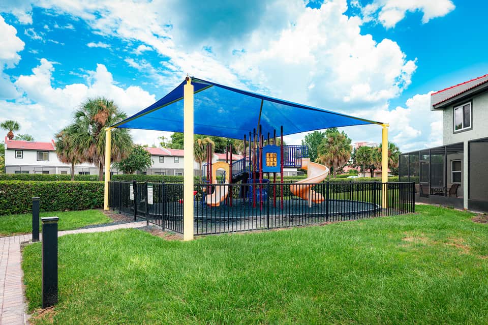 Playground with colorful equipment under blue shade canopy, surrounded by palm trees and green lawn