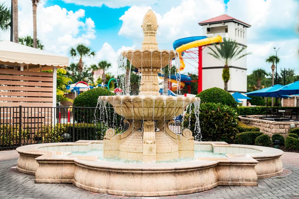 Ornate stone fountain in resort courtyard with palm trees, water slide, and blue umbrellas in background