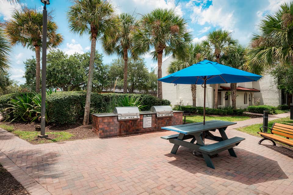 Outdoor patio with picnic table, blue umbrella, grill station, and palm trees