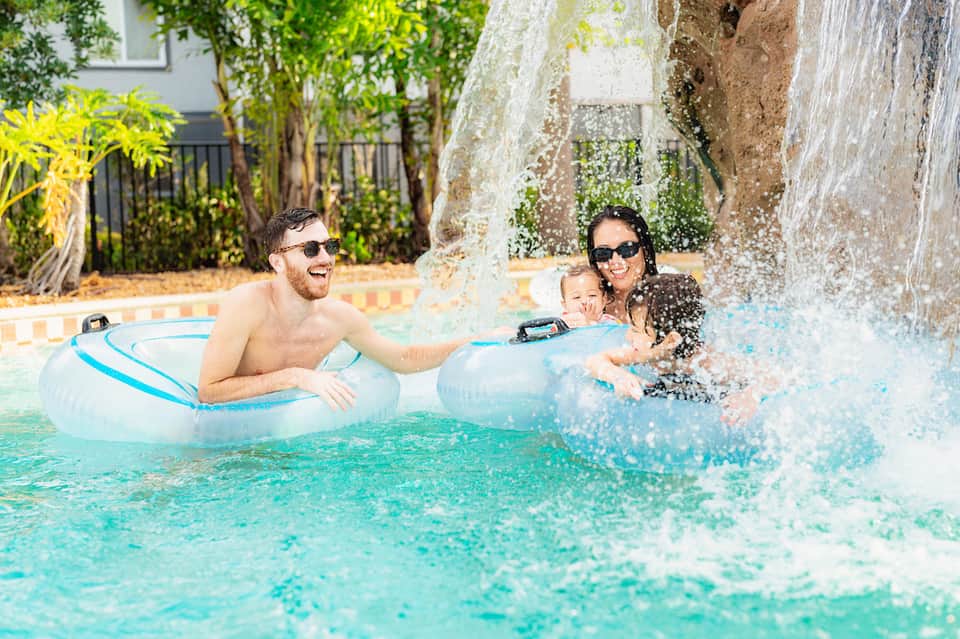 Family enjoying resort pool with waterfall feature, parents on float tubes with toddler in water