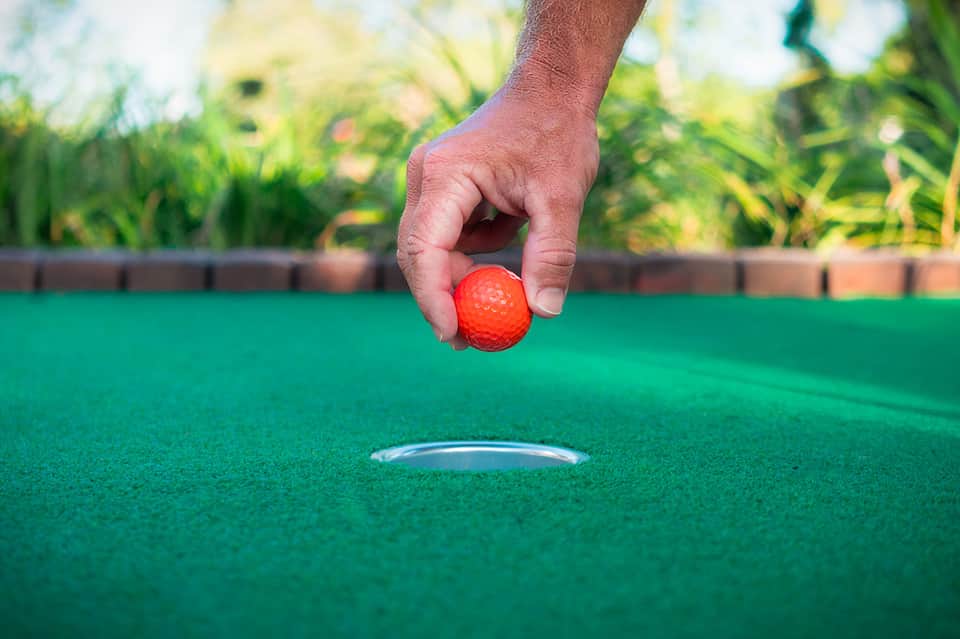 Hand holding red golf ball above hole on putting green with blurred garden background