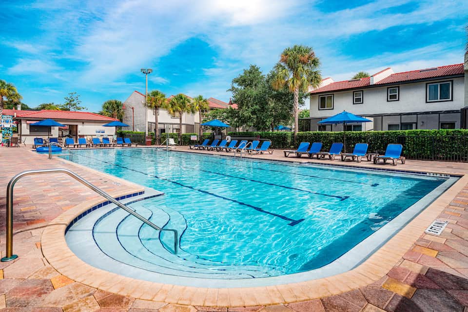 Olympic-sized swimming pool with blue lounge chairs, palm trees, and resort buildings under clear sky