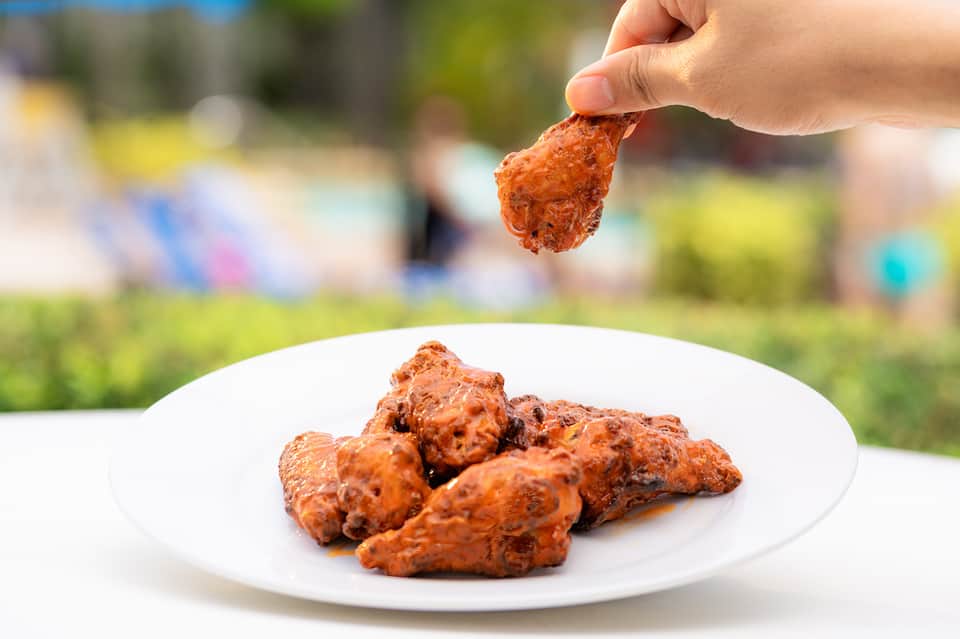 Hand holding crispy fried chicken wing over white plate at outdoor gathering