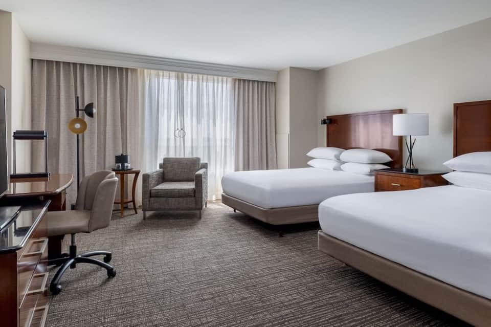 Hotel guest room with two beds, work desk, armchair, and balcony access