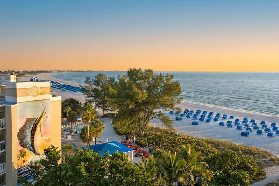 Beachfront resort at sunset with blue umbrellas on sand, ocean view, and illuminated building with tarpon mural