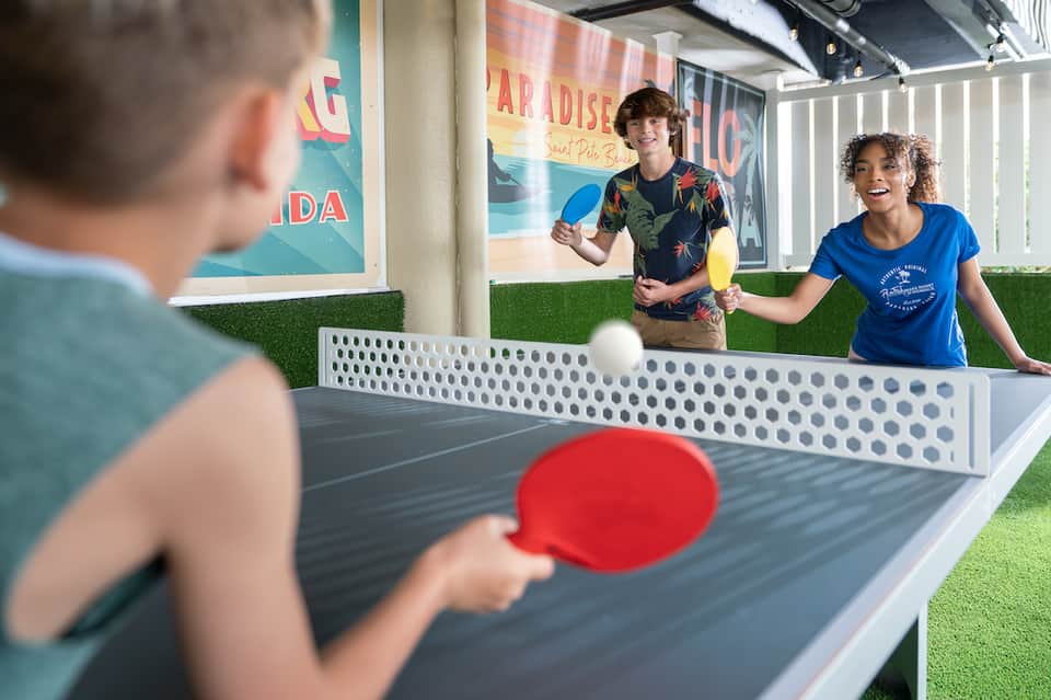 Children playing ping pong outdoors on artificial turf with tropical signage in background