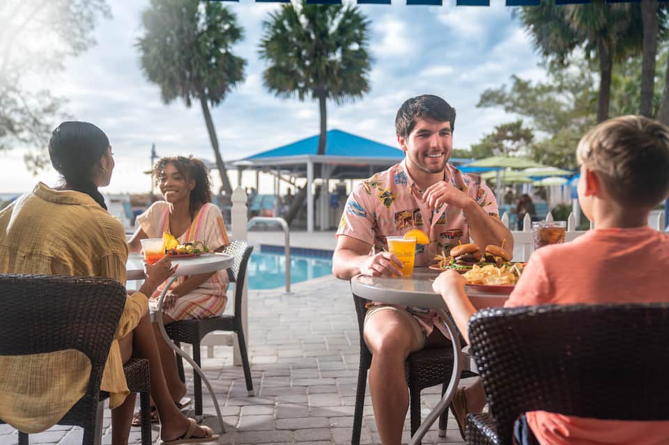 Guests dining poolside at umbrella-shaded tables with palm trees and blue cabana