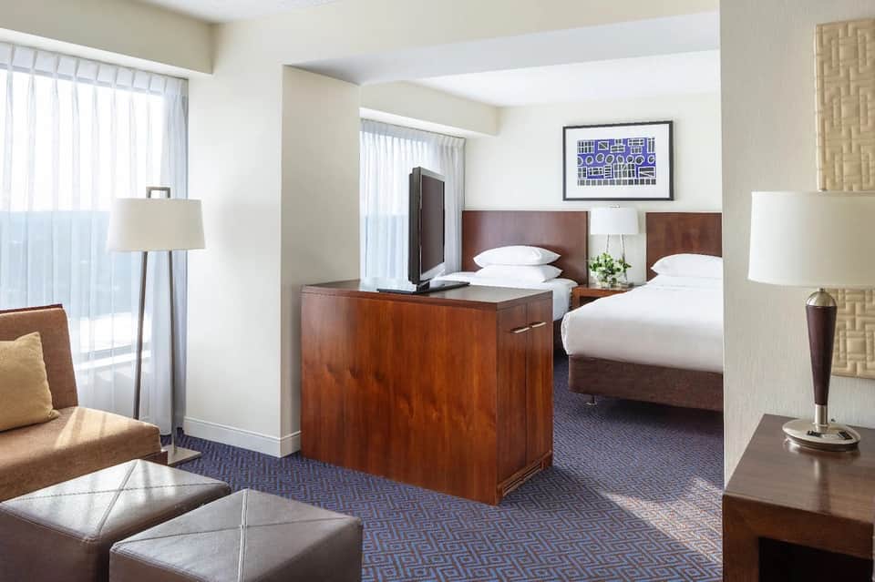 Hotel room with two queen beds, wooden headboards, white bedding, lounge seating, and bright windows