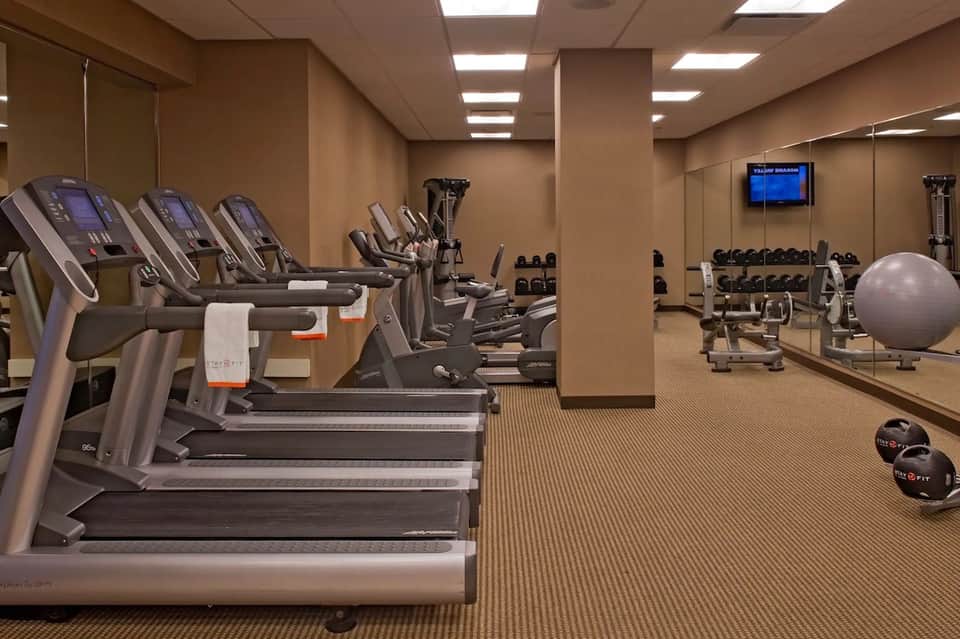 Fitness center with rows of treadmills, exercise bikes, dumbbells, stability balls, and wall-mounted TV