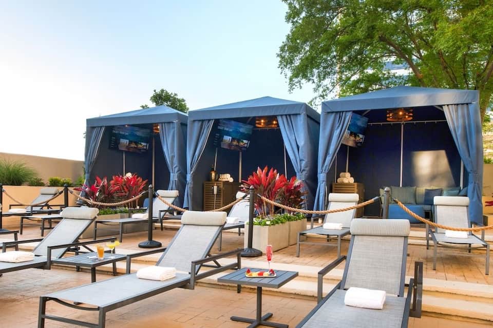 Luxury poolside cabanas with blue curtains, lounge chairs, and red flowering plants on terracotta patio
