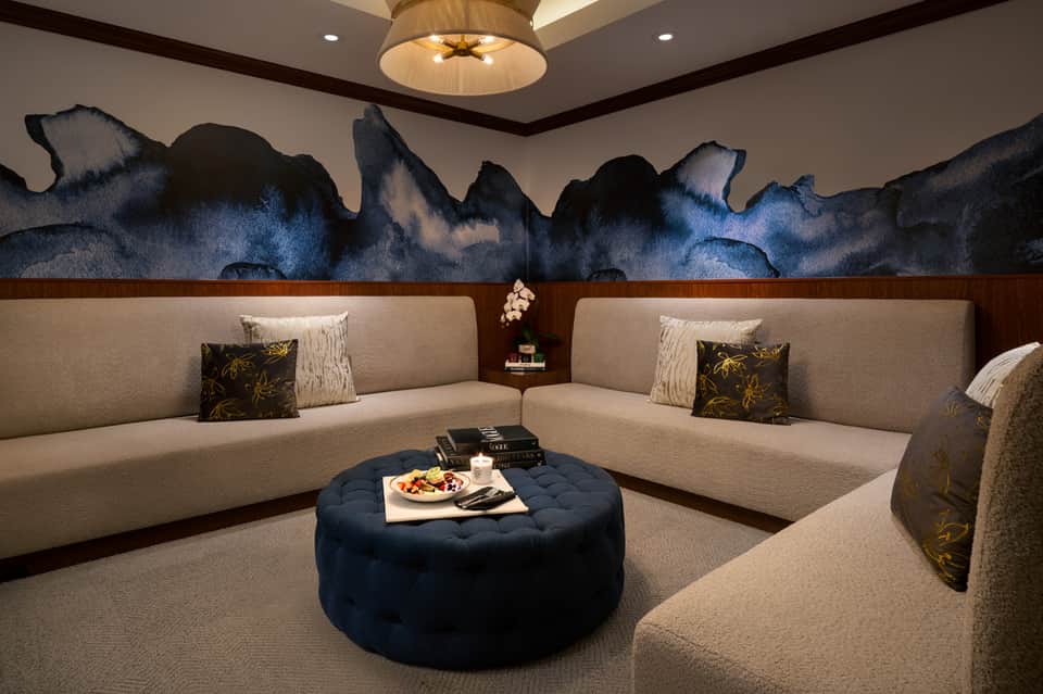 Luxury yacht lounge with L-shaped beige seating, navy ottoman, artistic wall mural, and round ceiling fixture
