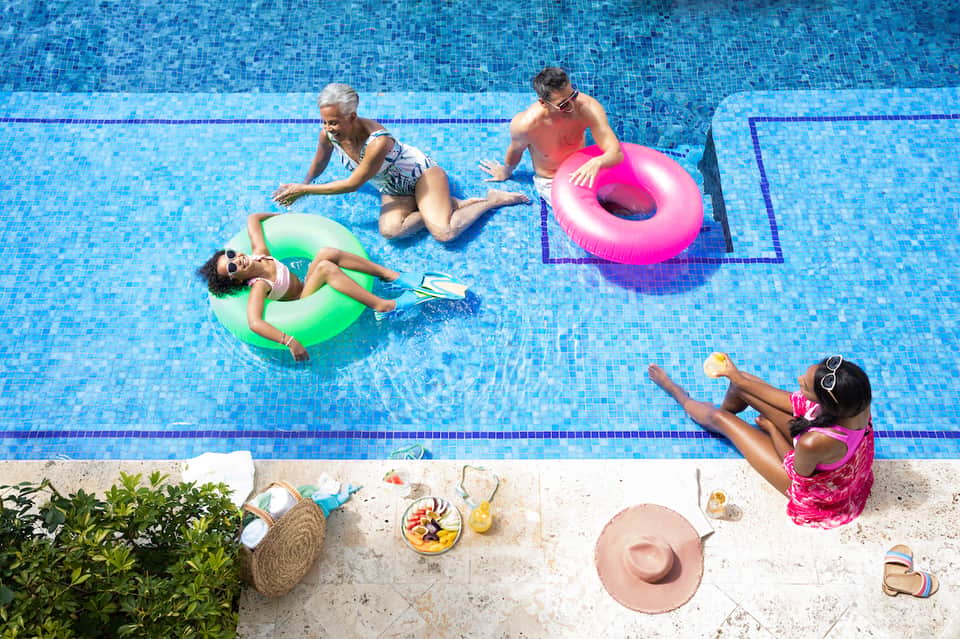Family in swimming pool with colorful inflatable floats on sunny day with poolside snacks and palm trees