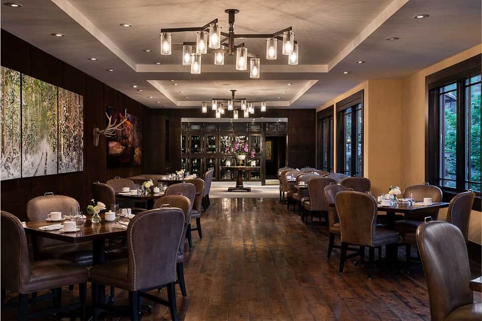 Upscale dining room with dark wood paneling, leather chairs, round tables, chandeliers, and bar area