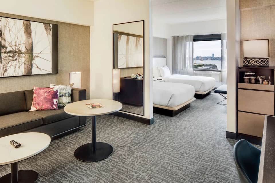 Modern hotel suite with living area, bedroom, and city waterfront view through floor-to-ceiling windows