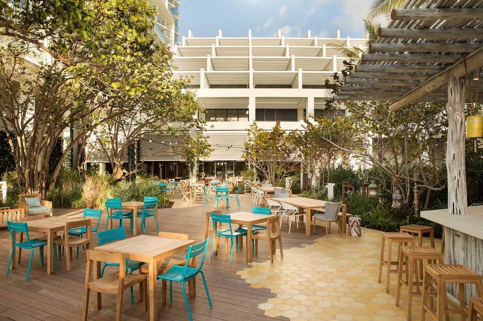 Outdoor courtyard dining area with turquoise chairs, wooden tables, and modern white architectural building overhead