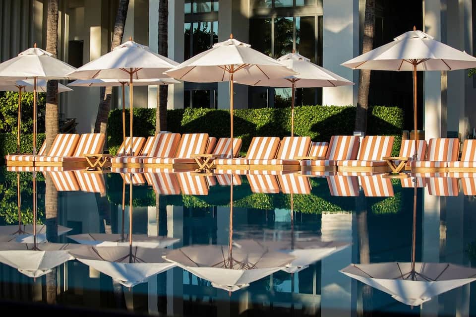 Poolside lounge area with striped orange and white chaise lounges, white umbrellas, and palm trees reflected in water