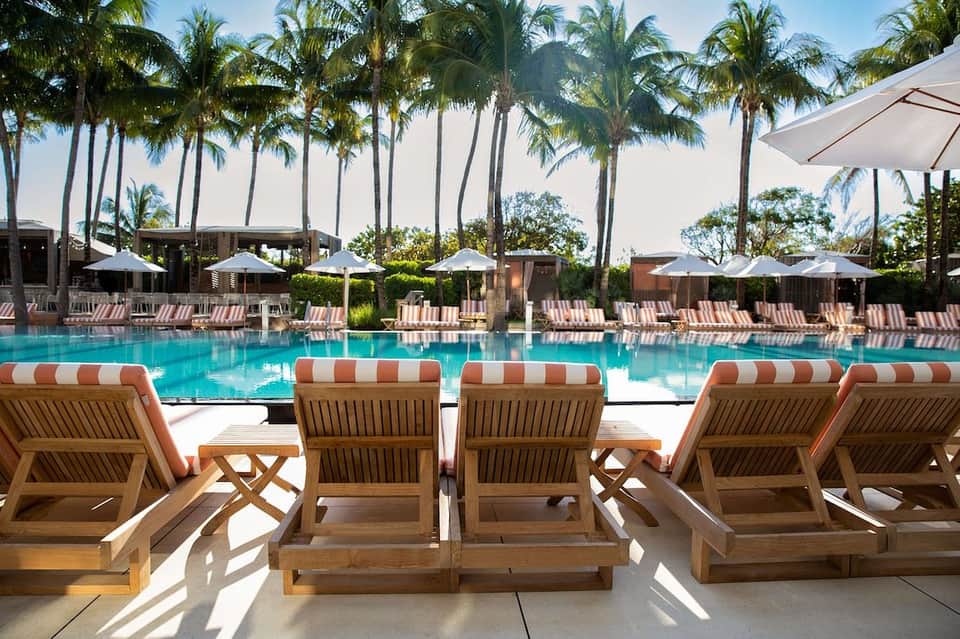 Resort pool deck with wooden lounge chairs, striped orange and white cushions, white umbrellas, and palm trees