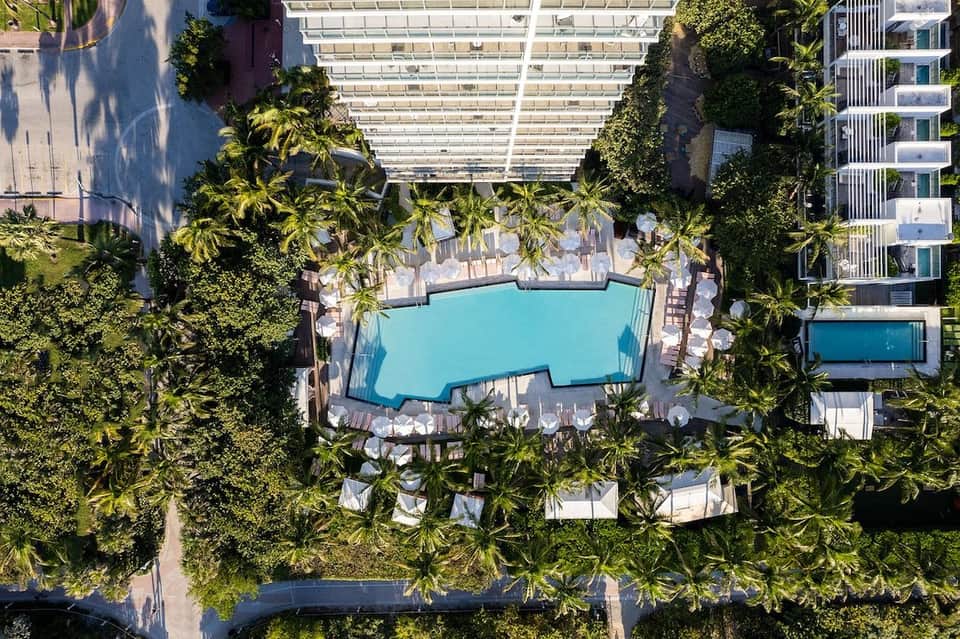 Aerial view of turquoise pool surrounded by palm trees, lounge chairs, and buildings with pergola shade structures