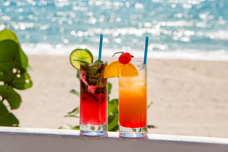 Two colorful tropical cocktails with lime and orange garnish on beach with ocean backdrop