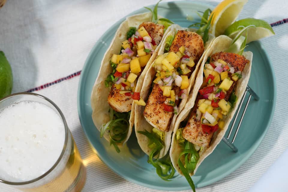 Three fish tacos with seasoned protein and pineapple salsa on light blue plate with lime wedges