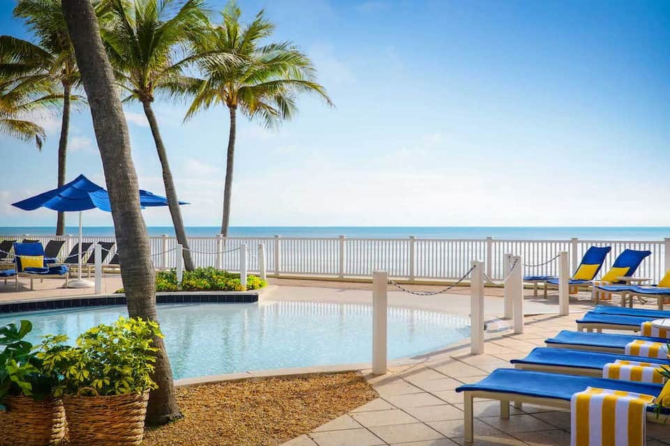 Beachfront resort pool area with blue umbrellas, lounge chairs, palm trees, and ocean view