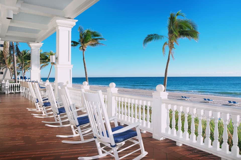 White rocking chairs on wooden deck overlooking beach with palm trees and clear ocean