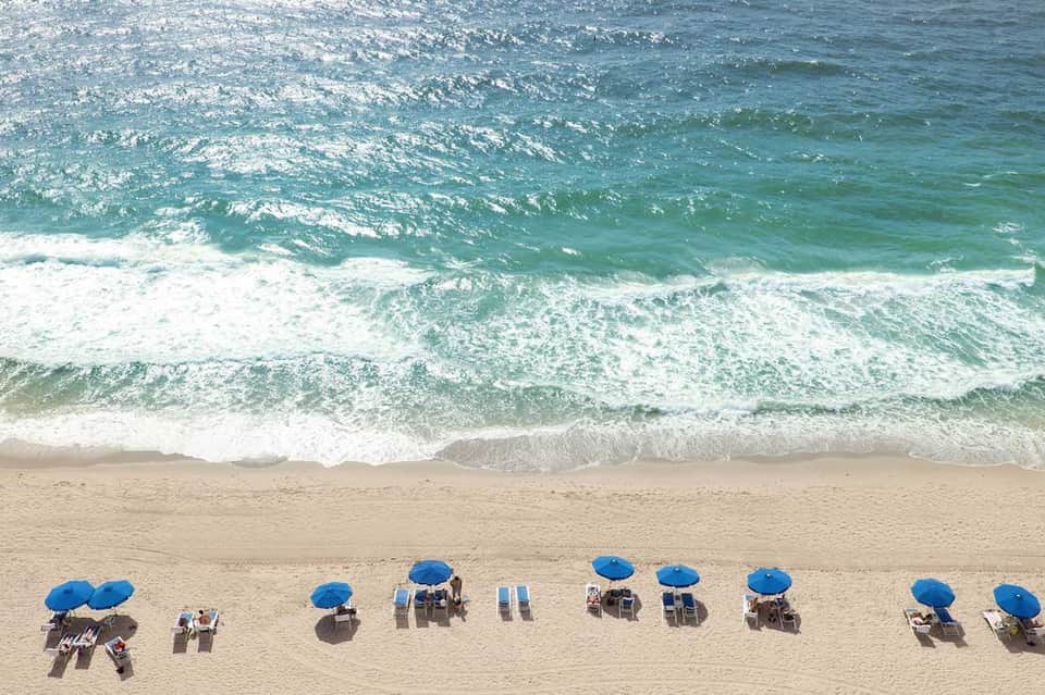 Aerial view of sandy beach with blue umbrellas and loungers lined up along calm turquoise ocean waves