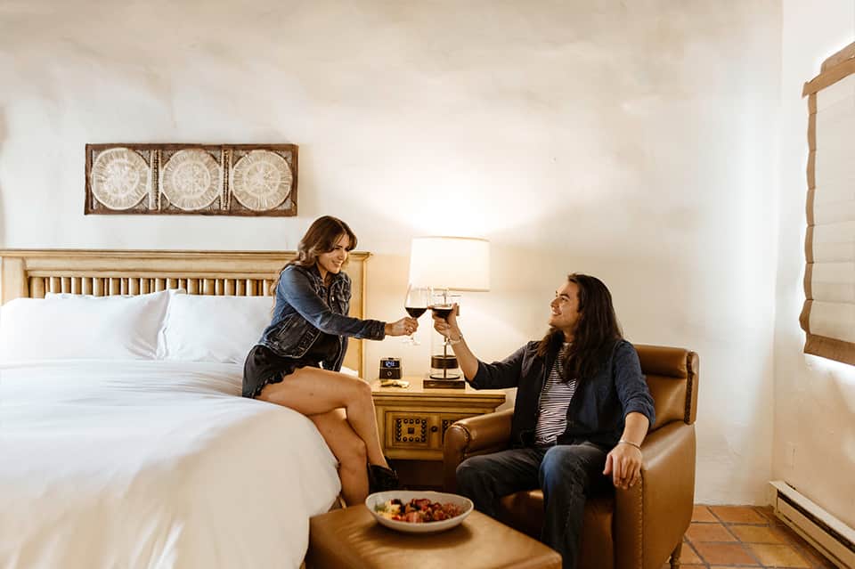 Two women toasting with wine glasses on a bed in a bright, minimalist hotel room