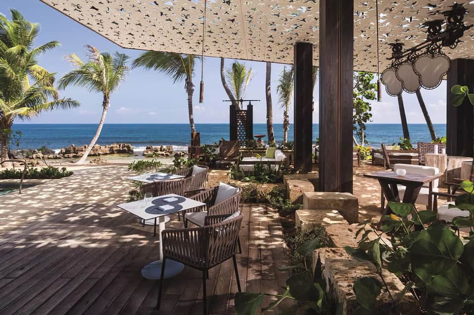 Beachfront dining pavilion with wooden tables, palm trees, and ocean views