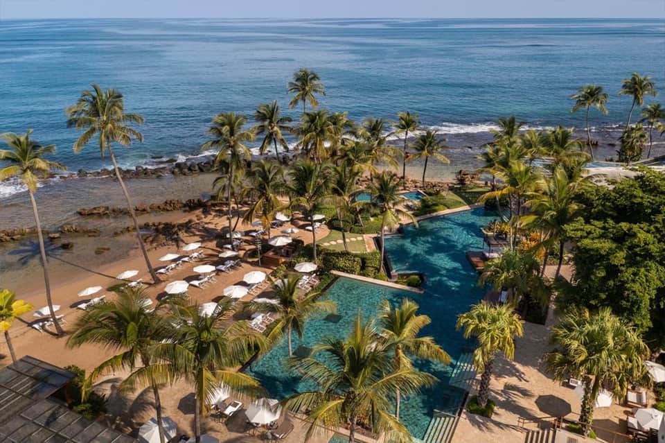 Tropical beachfront resort with multiple pools, white umbrellas, palm trees, and ocean views